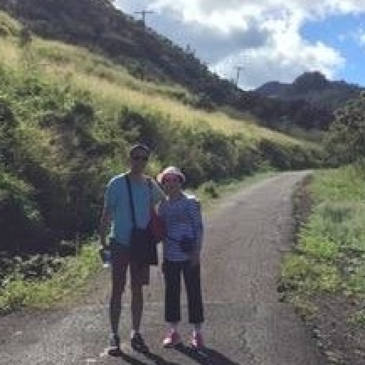 Two people stand on a paved path beside a grassy hillside under a partly cloudy sky.