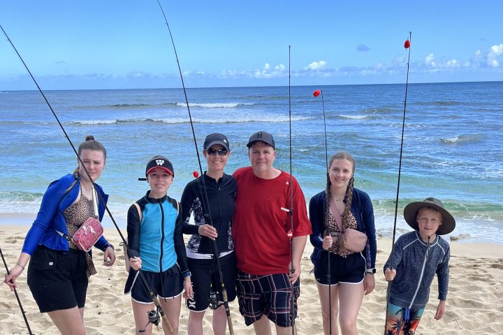 Group of six people with fishing rods standing on a sandy beach.