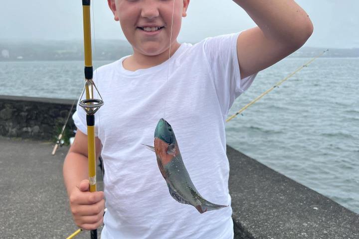 Boy holding a small fish with a fishing rod by a waterfront on a cloudy day.