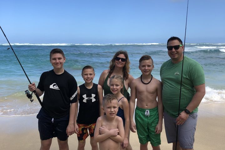Family of six posing on a beach with fishing poles under a blue sky.
