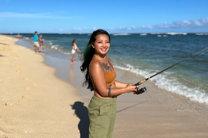 Woman fishing on a sandy beach next to the ocean under a clear blue sky.