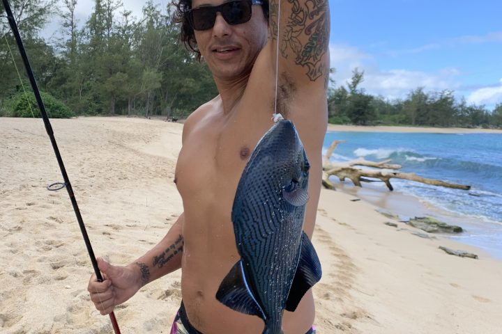 Person holding a fishing rod and fish on a beach, wearing floral shorts and sunglasses.