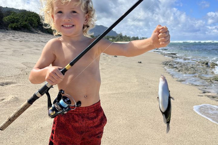 Smiling child in red shorts holds a fishing pole with a fish on a sunny beach.