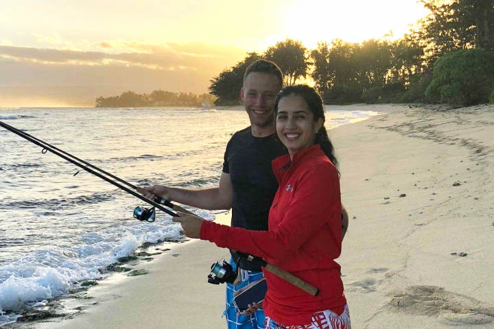 Two people fishing on a beach at sunset, with waves and trees in the background.