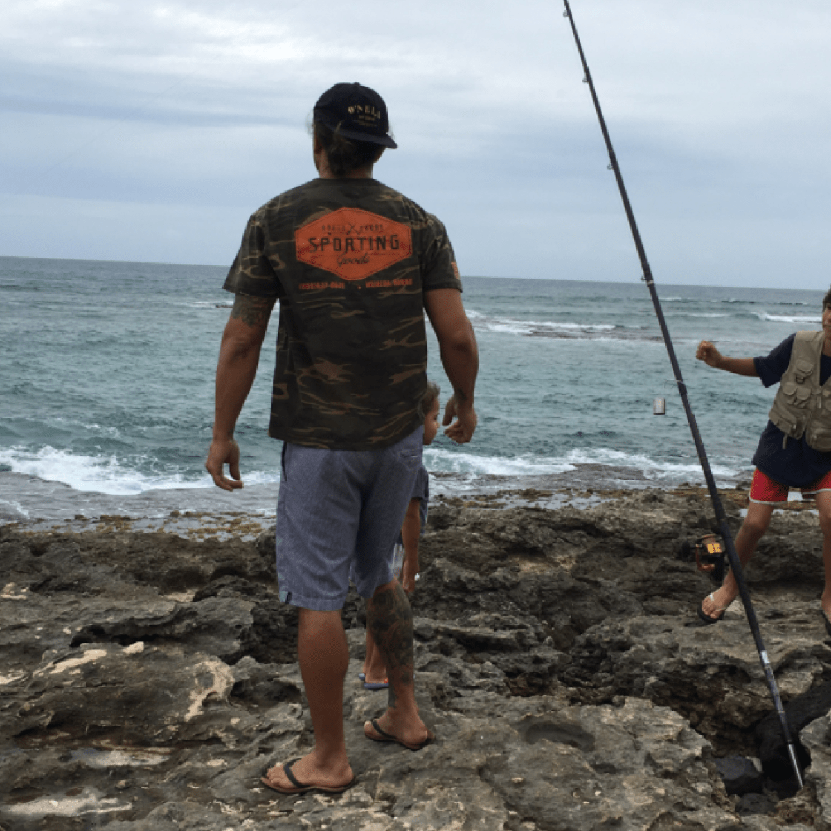 Two people fishing on rocky shore, cloudy sky above ocean waves.